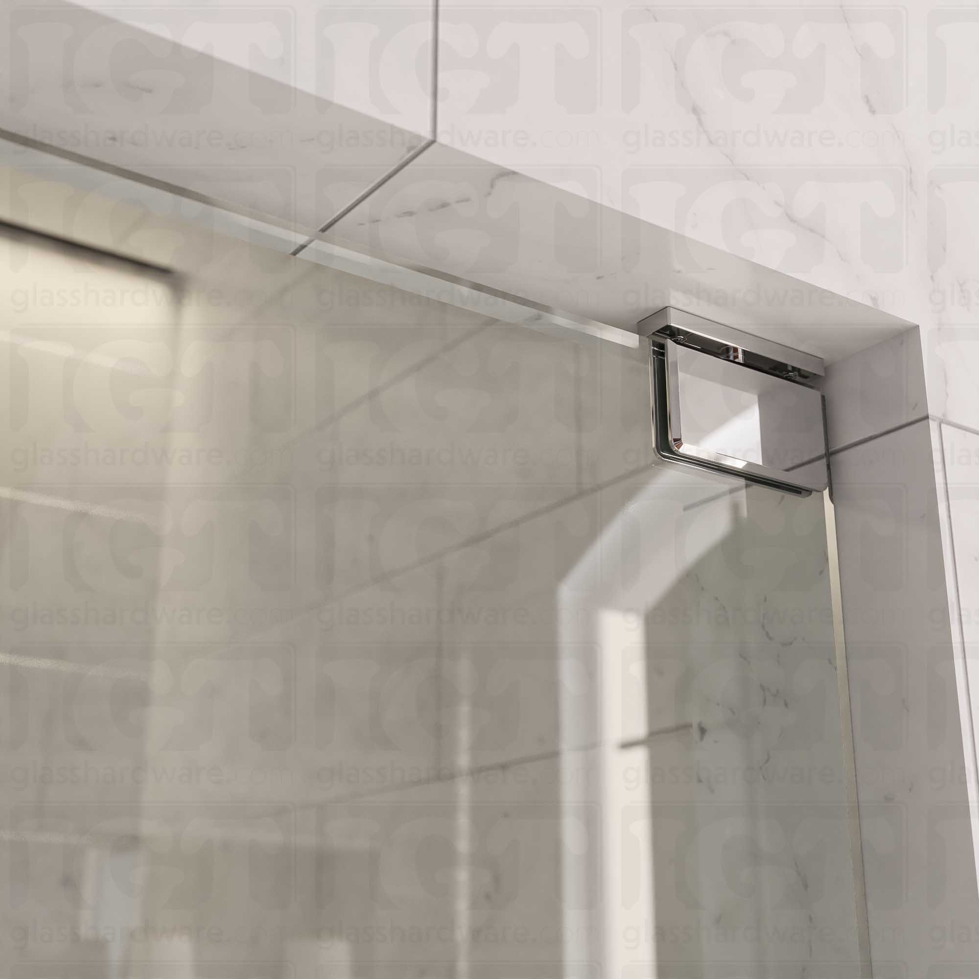 Close-up of the Top and Bottom Bilboa Pivot Hinge mounted at the top of the glass shower door, securing it to the tiled wall. The hinge has soft rounded corners and beveled edges, making it blend into the elegant bathroom setting. Chrome Polished.