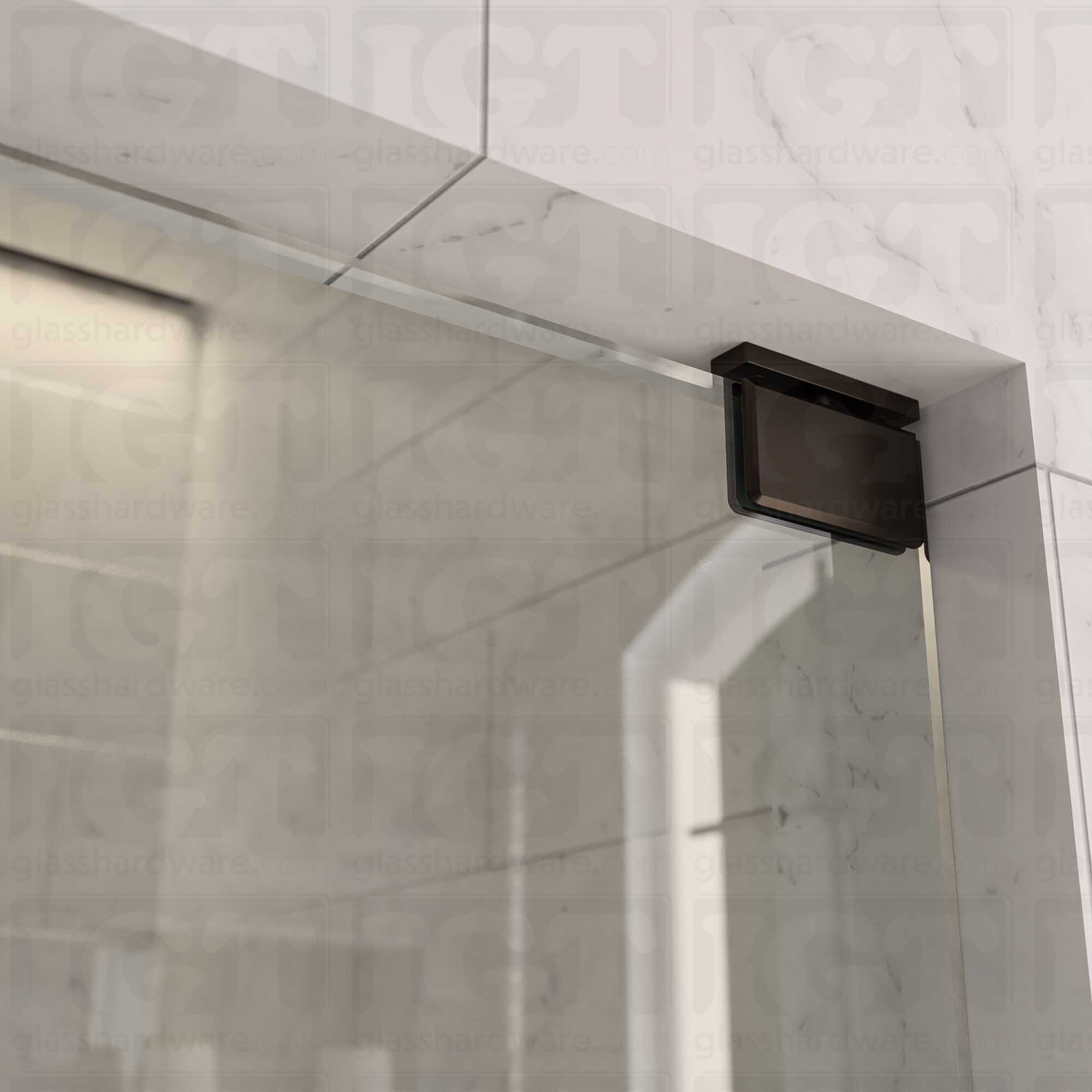 Close-up of the Top and Bottom Bilboa Pivot Hinge mounted at the top of the glass shower door, securing it to the tiled wall. The hinge has soft rounded corners and beveled edges, making it blend into the elegant bathroom setting. Oil Rubbed Bronze.