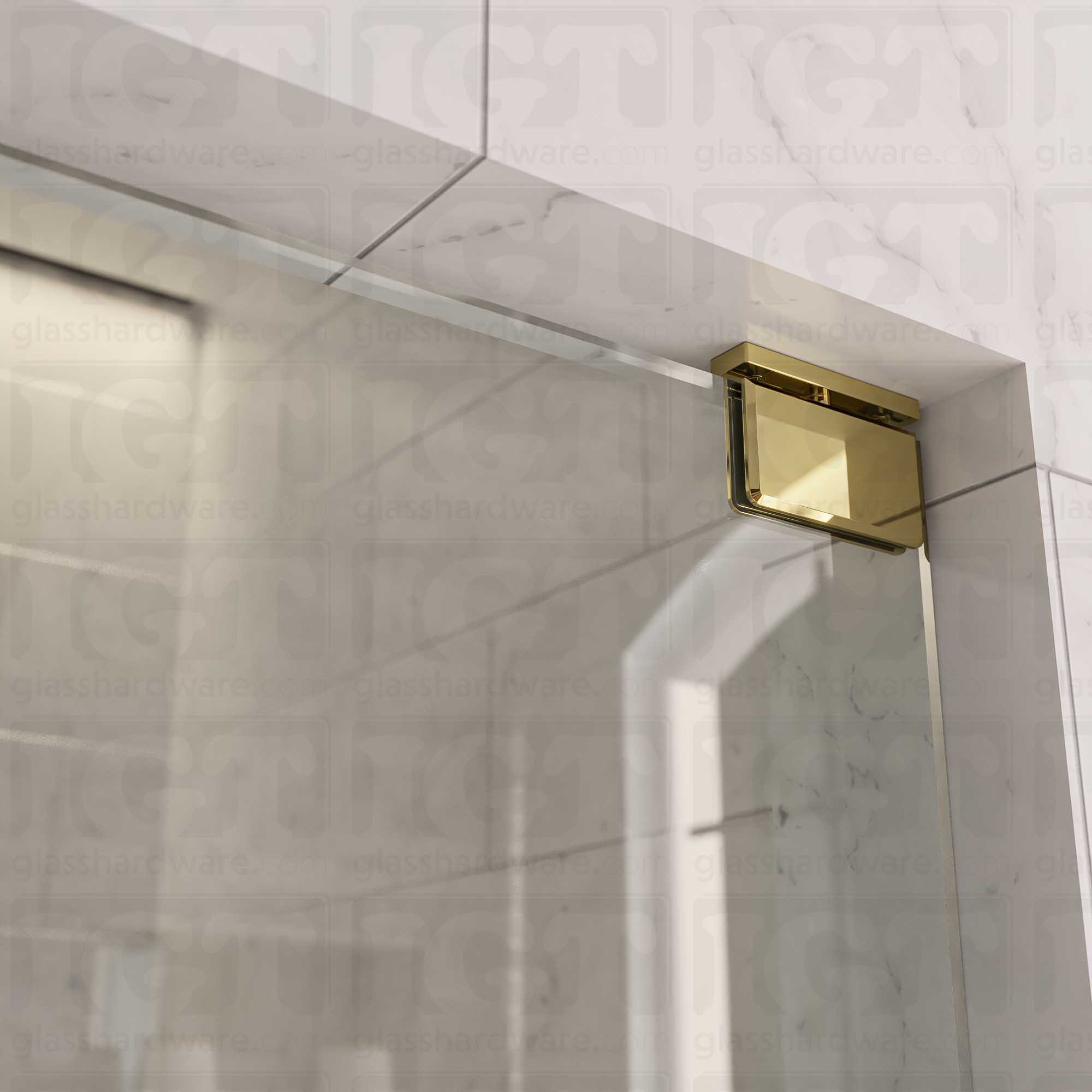 Close-up of the Top and Bottom Bilboa Pivot Hinge mounted at the top of the glass shower door, securing it to the tiled wall. The hinge has soft rounded corners and beveled edges, making it blend into the elegant bathroom setting. Polished Brass.