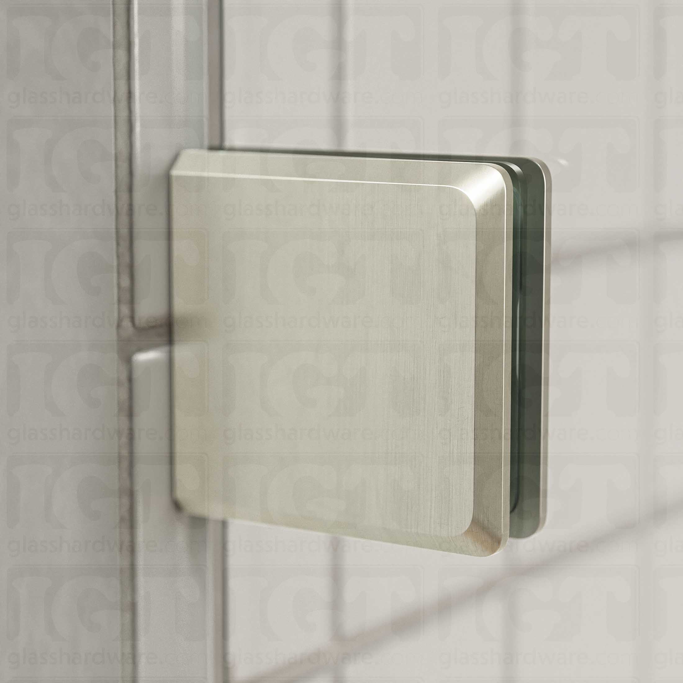 Close-up view of a Wall-to-Glass Bilboa Clamp mounted to the shower's white tile wall. Natural light from the shower window reflects off the clamp's beveled edges. Brushed Nickel.