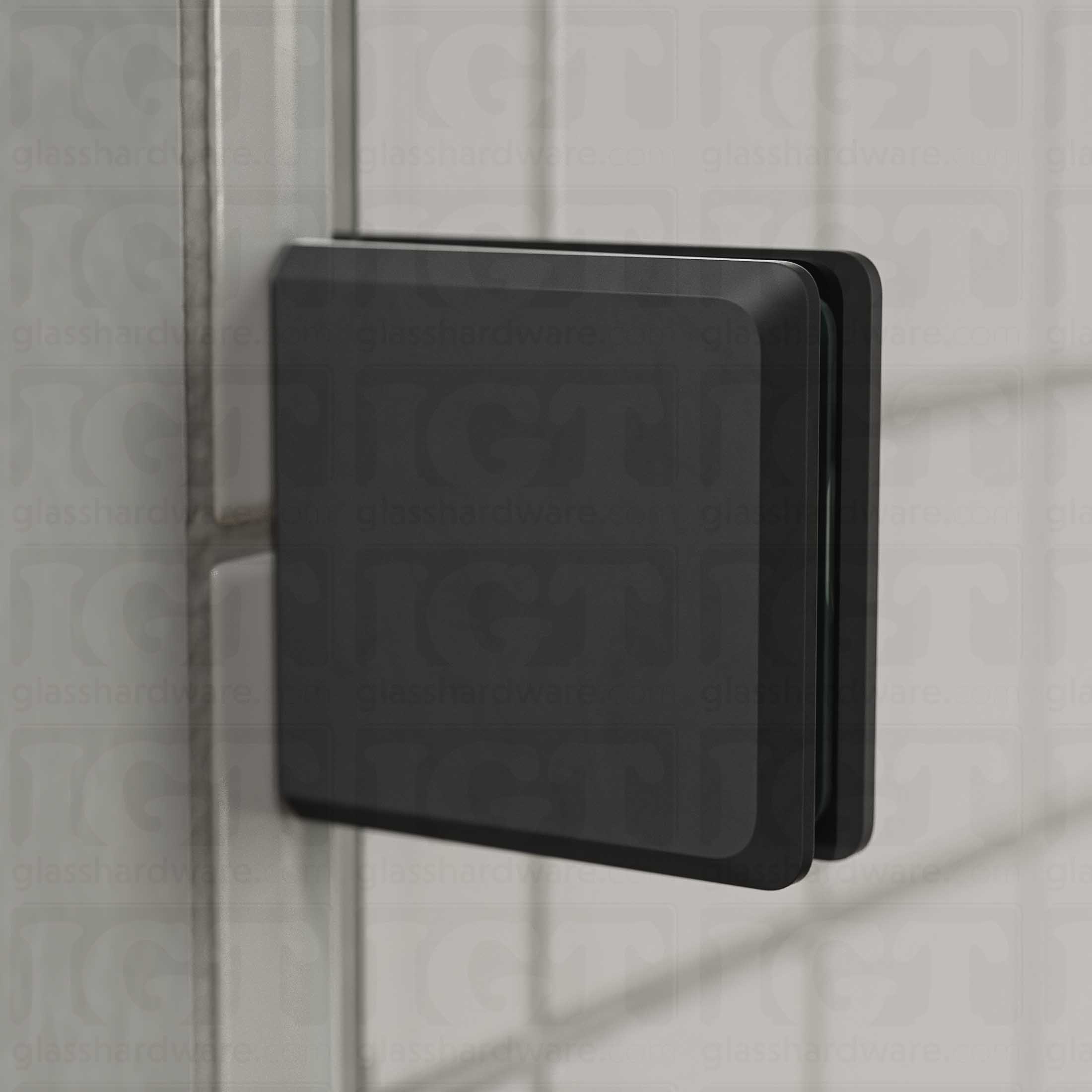 Close-up view of a Wall-to-Glass Bilboa Clamp mounted to the shower's white tile wall. Natural light from the shower window reflects off the clamp's beveled edges. Matte Black.