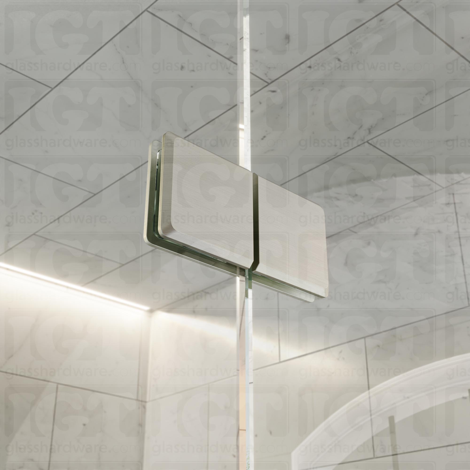 A close-up view of the Glass-to-Glass 180 Degree Bilboa Clamps (Split Face), installed on a frameless glass shower enclosure. Natural light travels along the clamp's sleek beveled edges and adds dimension to its overall design. Brushed Nickel.