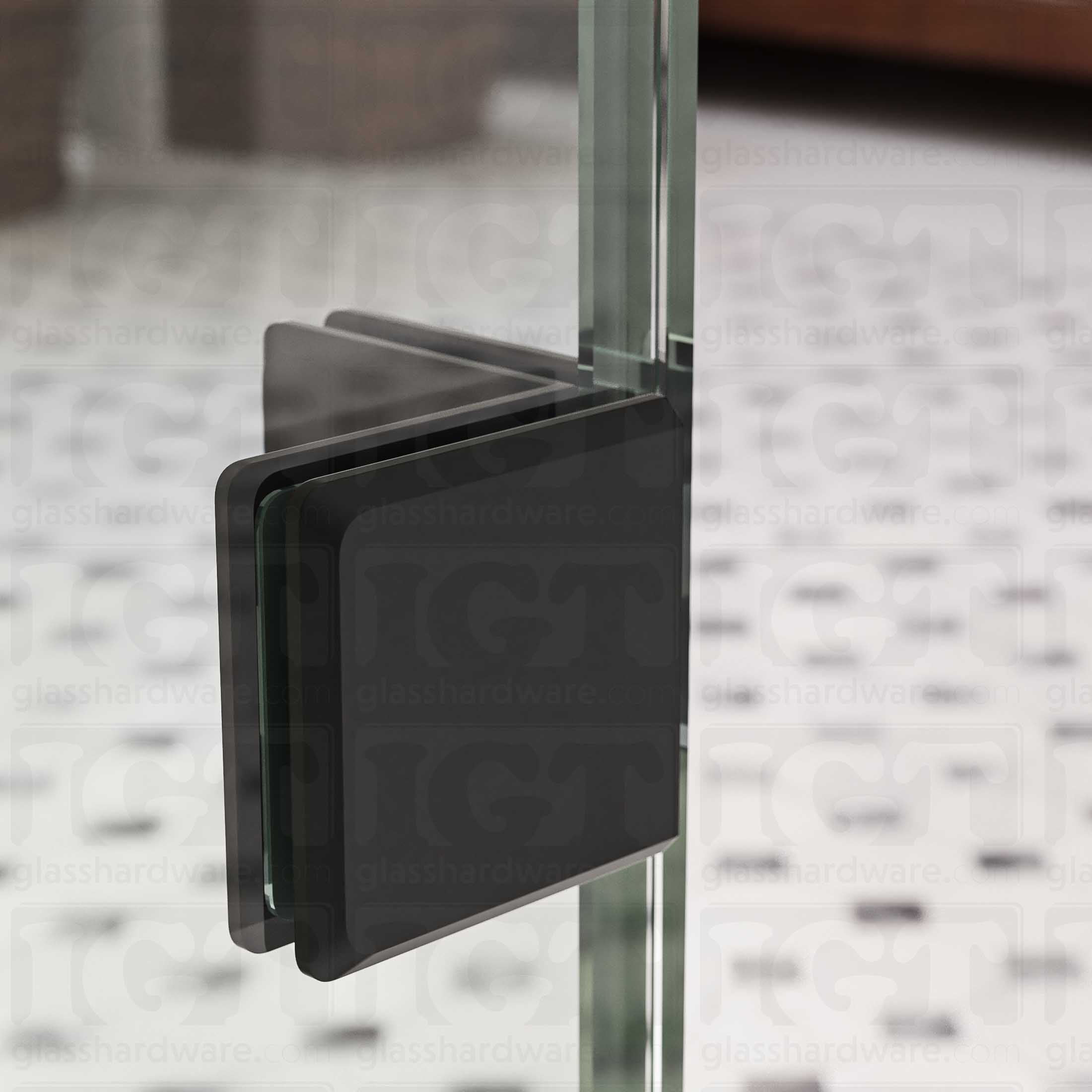 A close-up view of the Glass-to-Glass 90 Degree Bilboa Clamp, installed on a frameless glass shower enclosure. Natural light reflects along the clamp's sleek beveled edges and adds dimension to its overall design. Matte Black.