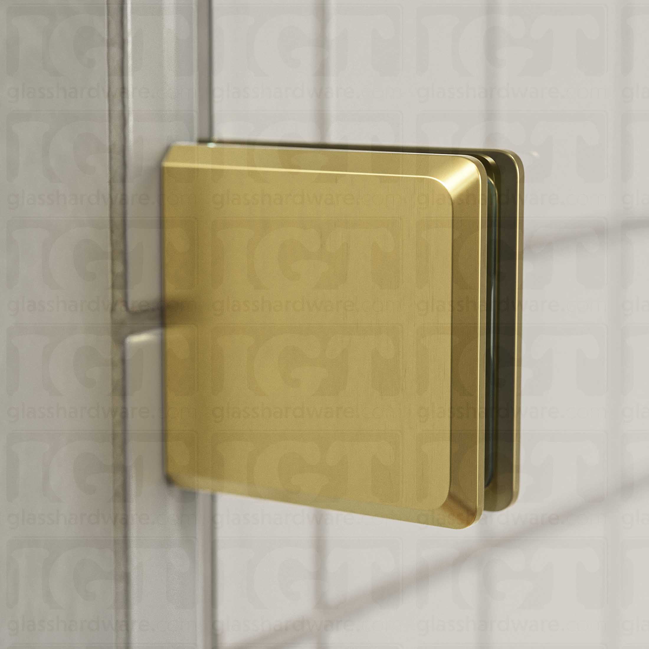 Close-up view of a Wall-to-Glass Bilboa Clamp mounted to the shower's white tile wall. Natural light from the shower window reflects off the clamp's beveled edges. Gold Brushed.