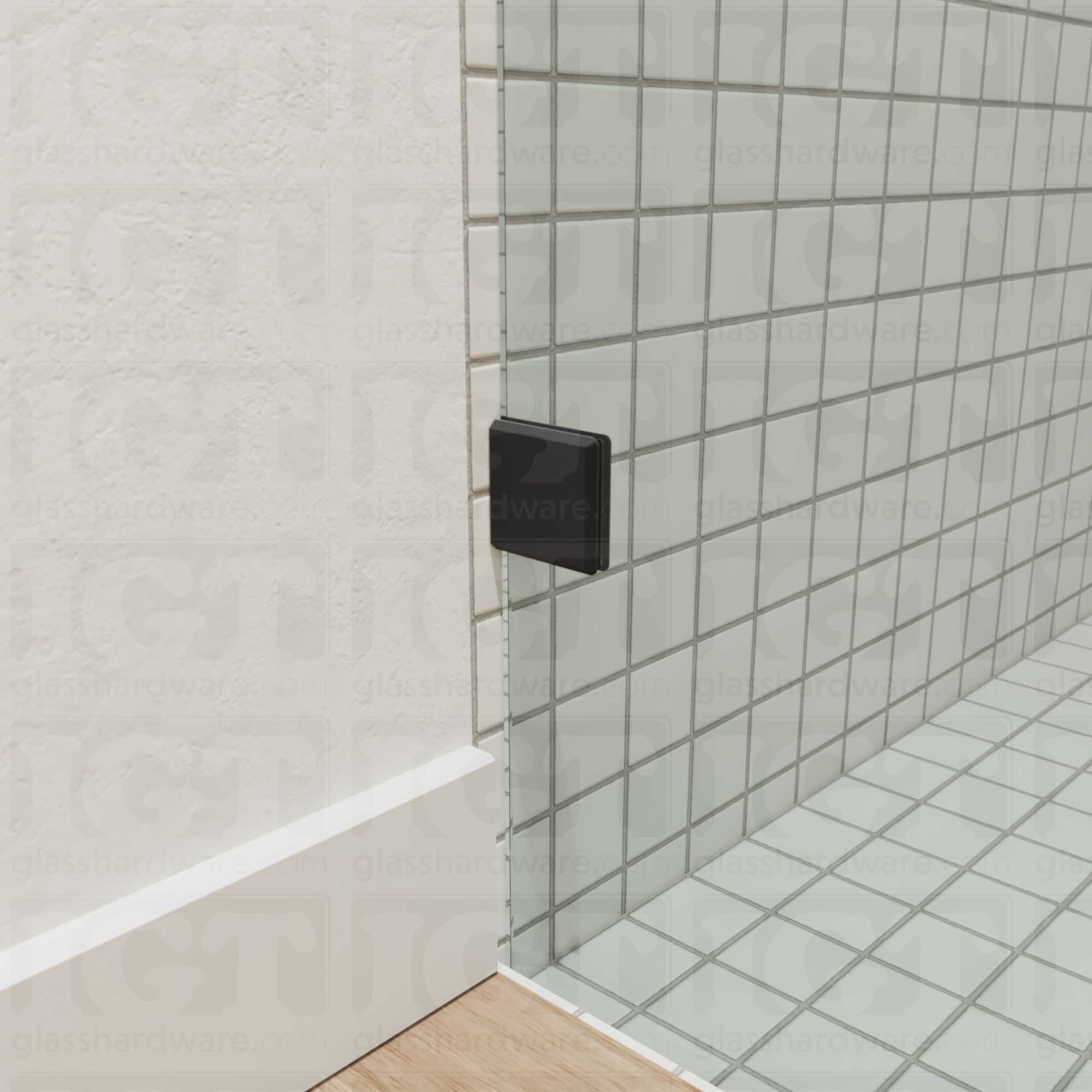 A close-up view of the bottom corner of the glass shower enclosure, showing an Oversized Wall-to-Glass Bilboa Clamp securing the panel to the wall. Matte Black.