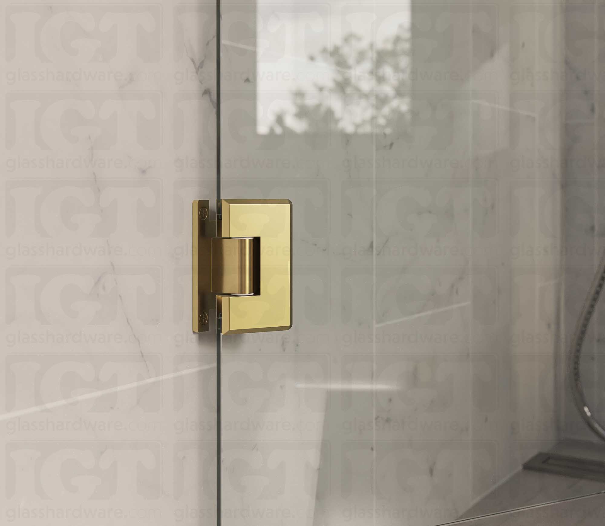 A close up view of the Heavy Duty Wall-to-Glass “H” Back Bilboa Hinge installed on a frameless glass shower door. The bathroom's natural lighting reflects along the hinge's sleek beveled edges, adding dimension to the overall design. Gold Brushed.